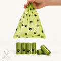 A hand holds a translucent light green plastic bag printed with black paw prints, shown above several matching rolled bags on a white studio background. A PET MANYA logo appears in the lower left corner.