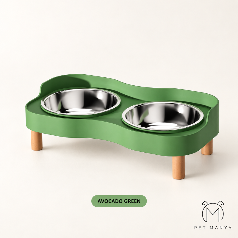 A green elevated double pet feeder with stainless steel bowls and wooden legs, shown on a clean off-white background with a label reading 'AVOCADO GREEN' and the Pet Manya logo.