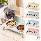 A gray-and-white cat is eating from a pastel green elevated dual-bowl pet feeder with stainless steel bowls, shown in a warm indoor setting. Smaller views of the same feeder in yellow, mint, blue, and pink appear on the right, along with the brand name PET MANYA.