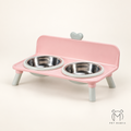 Pink elevated pet feeder and waterer with two stainless steel bowls, shown on a clean cream background. The feeder has light gray legs and a small gray heart detail on the backrest.