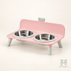 Pink elevated pet feeder and waterer with two stainless steel bowls, shown on a clean cream background. The feeder has light gray legs and a small gray heart detail on the backrest.