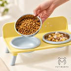 A yellow elevated pet feeding station with two bowls, shown with a hand pouring dry cat food into one bowl and wet cubed food into the other. The brand name 'PET MANYA' is visible in the bottom right corner.