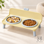 A yellow elevated pet feeder with two bowls, one filled with dry kibble and the other with cubed wet food, set in a softly lit indoor environment. The brand logo 'PET MANYA' is visible in the bottom right corner.