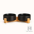 A pair of matte black pet bowls on a light natural wood base, photographed against a clean off-white background with the brand name 'PET MANYA' in the bottom right corner.