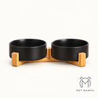 A pair of matte black pet bowls on a light natural wood base, photographed against a clean off-white background with the brand name 'PET MANYA' in the bottom right corner.