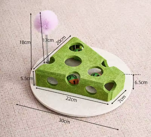 A green cheese-shaped cat toy with holes, mounted on a round white base, with a pink pom-pom stick on a spring. The image includes multiple black dimension lines and text labels showing measurements of the product and its parts on a beige textured background.