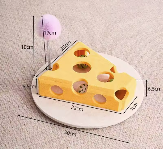 A cheese-shaped cat toy with a spring-mounted purple licker tail, shown on a round wooden base with overlaid dimension labels and arrows on a neutral fabric background.