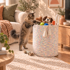 A fluffy tabby cat stands on its hind legs and places its paws on a woven pastel laundry basket filled with toys, in a bright, sunlit living room.